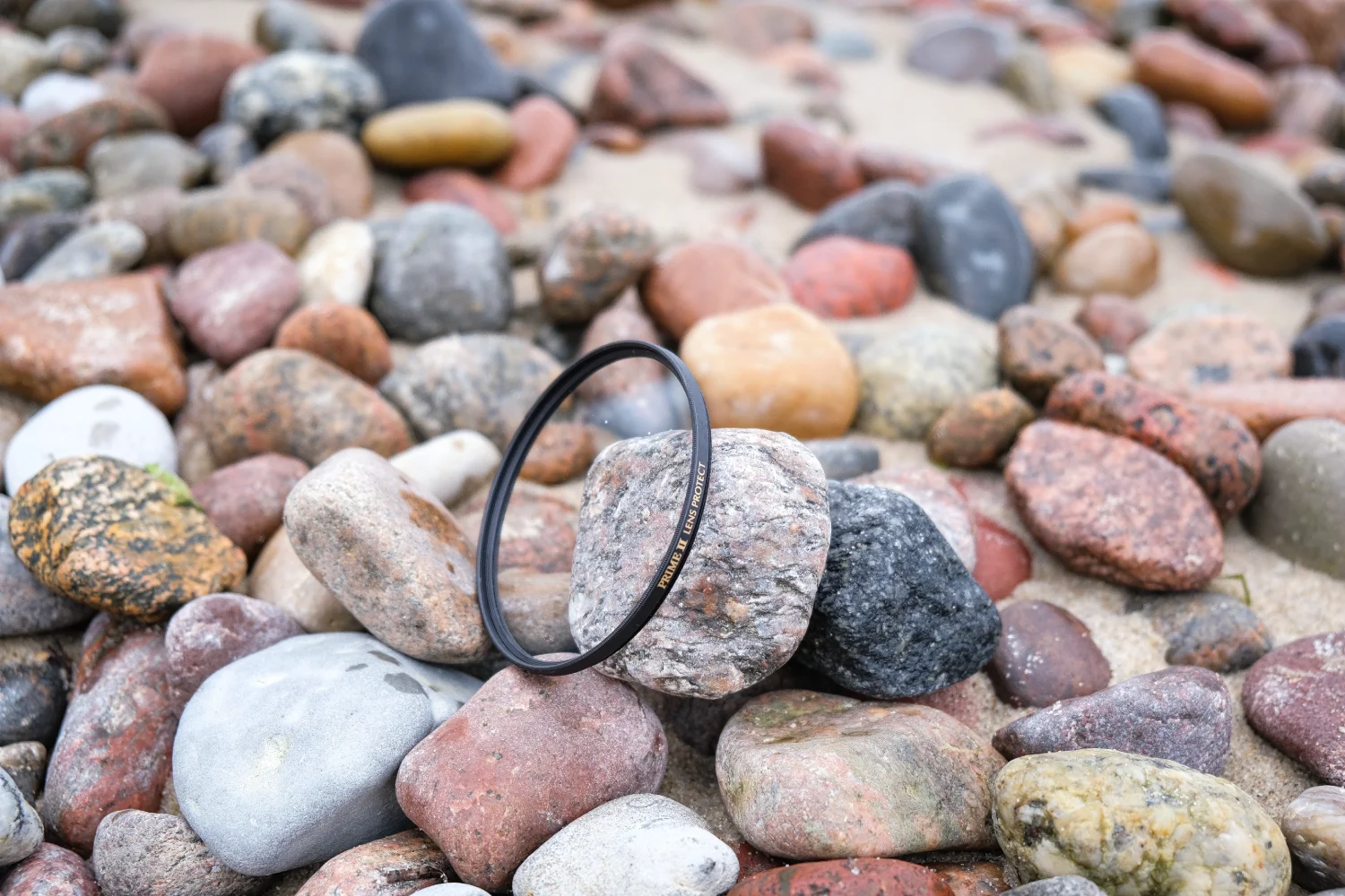 Marumi PRIME II Lens Protect – ochrona obiektywu w nowym wydaniu zdjęcie przedstawiające filtr Marumi Prime II Lens Protect leżący na kamieniach na brzegu nadmorskim z widocznym napisem PRIME II LENS PROTECT na oprawce filtra